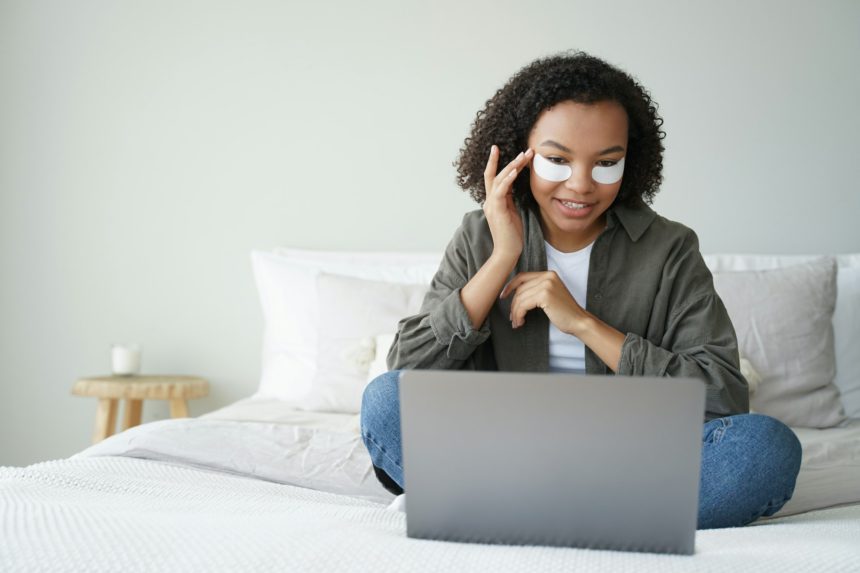 African american girl applying under eye patches sitting at laptop on bed. Skincare beauty routine